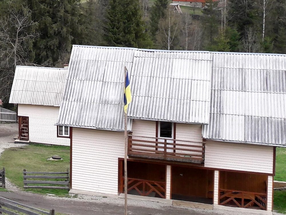 Світлий двоповерховий будинок з балконом і критою терасою внизу, прапор України на щоглі, за будинком смереки й ліс